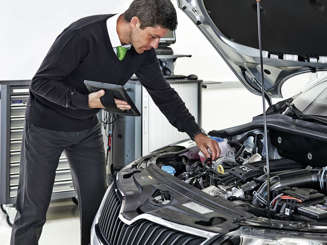 Mann in einer Werkstatt prüft den Motorraum eines Autos mit geöffnetem Fahrzeugdeckel und hält dabei ein Tablet zur Fahrzeugdiagnose.